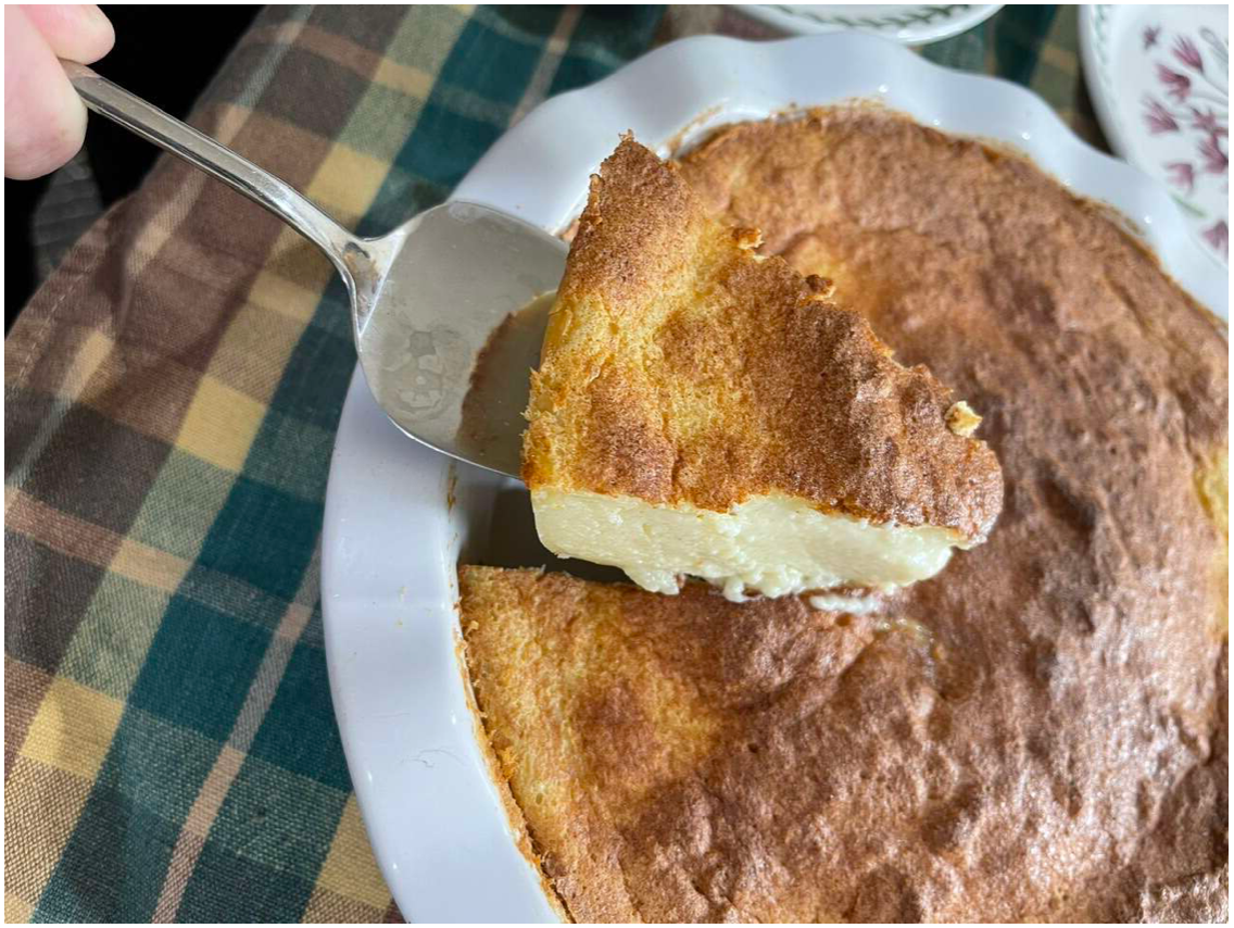 A slice of baked milk tart being lifted out of a pie dish with a cake slice showing a golden-brown top and light cream inside.